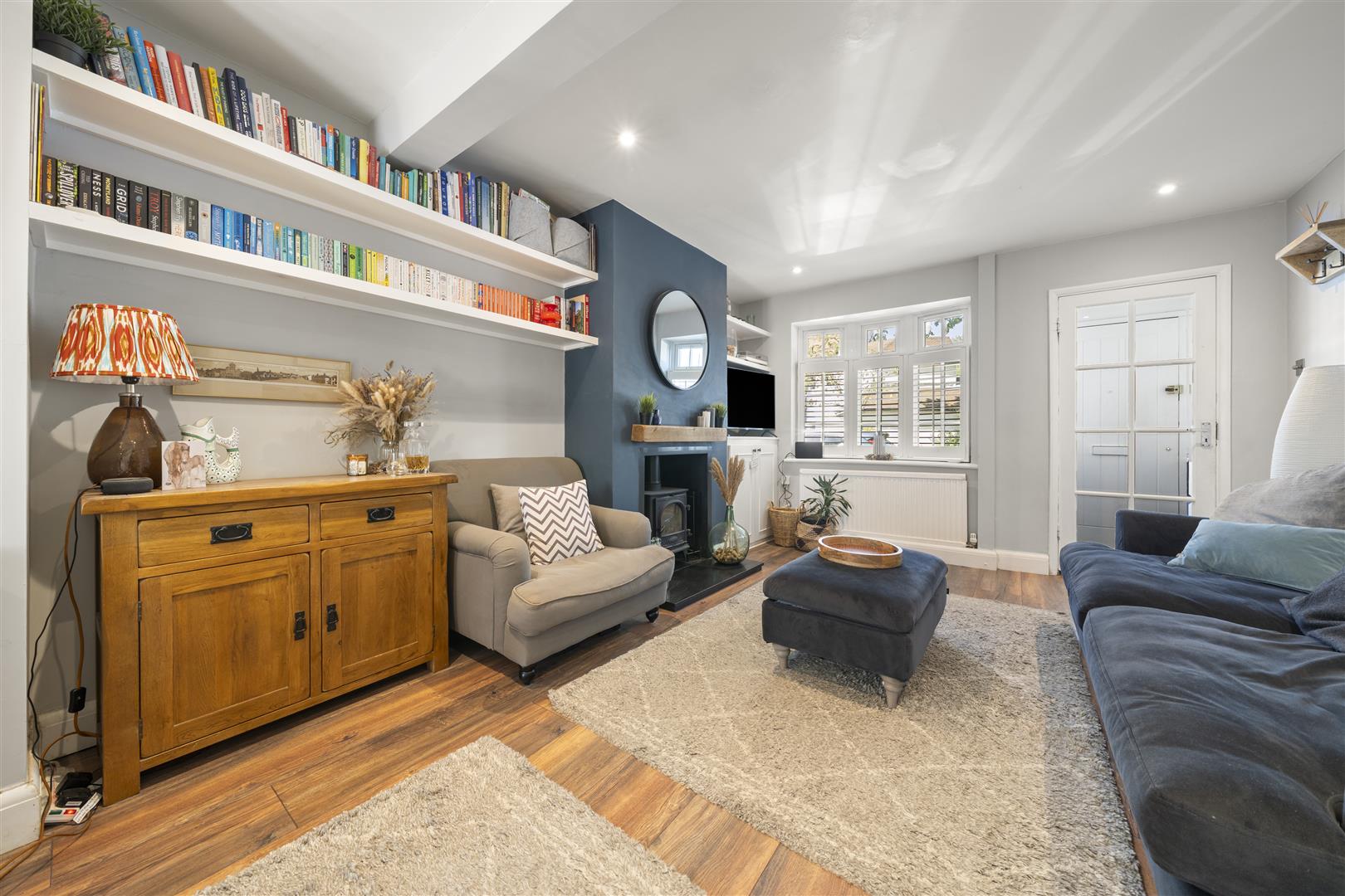 Cozy living room with tall white bookshelves full of colorful books, a beige armchair, and a wooden sideboard near a fireplace on a blue accent wall.