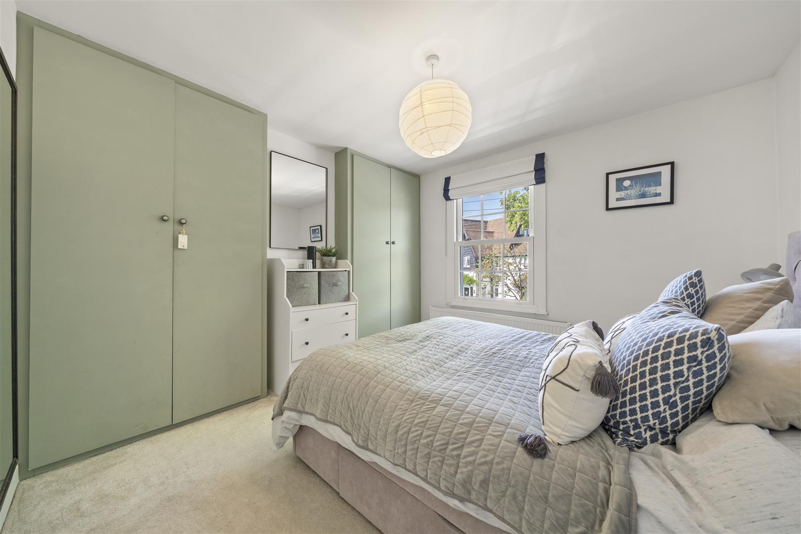 Bright bedroom with sage green wardrobes, a white dresser with fabric baskets, and a bed in a gray quilt with blue pillows.