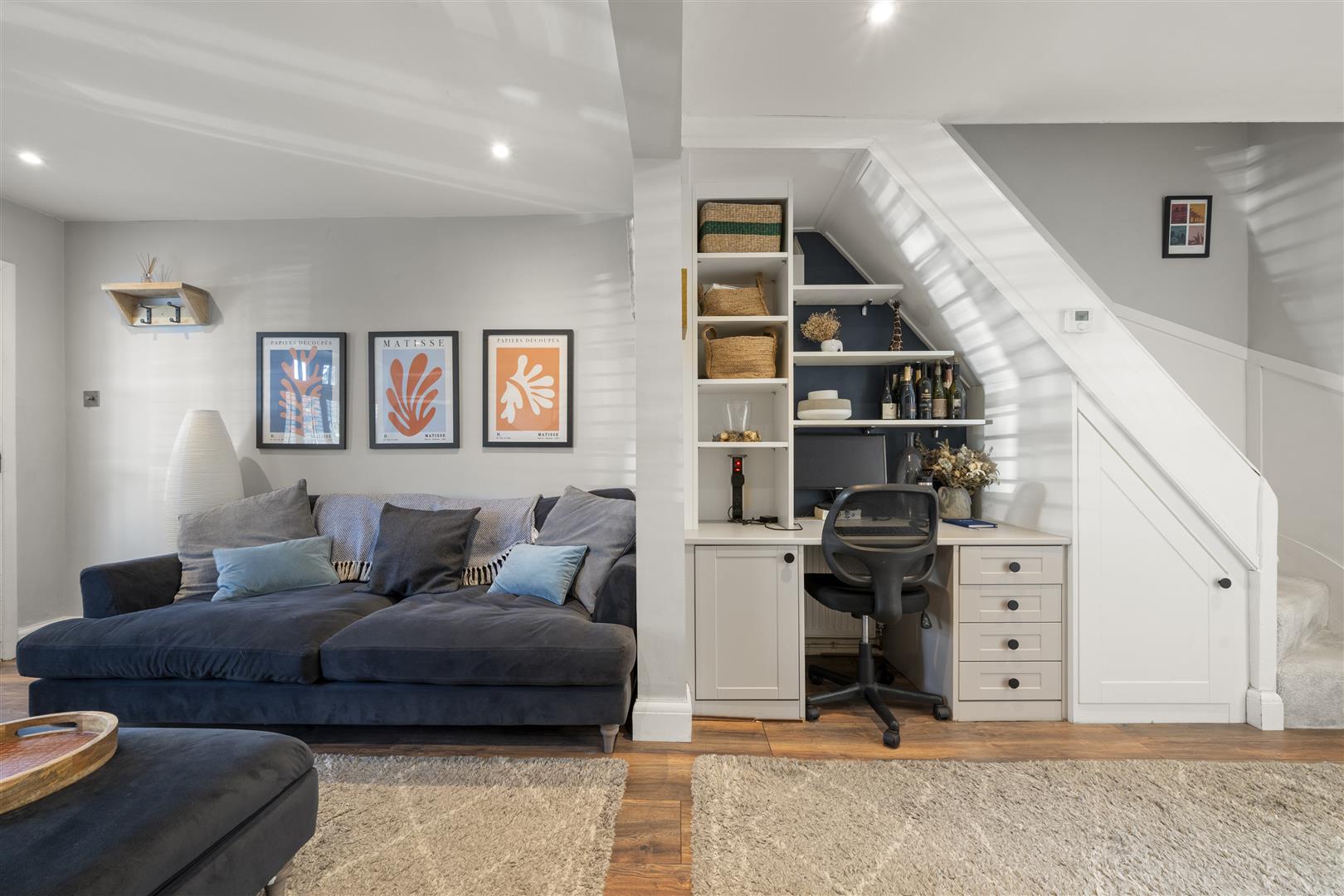 Cozy living area with a dark blue sectional, gray cushions, and framed art on a light gray wall; a white desk and shelves under a staircase.