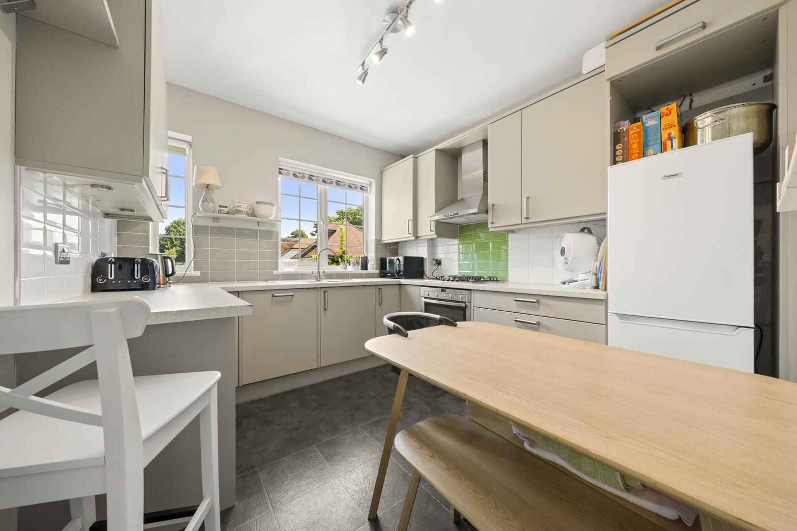 Bright modern kitchen with light gray cabinets, white appliances, and a wooden dining table in the foreground under a track light fixture.