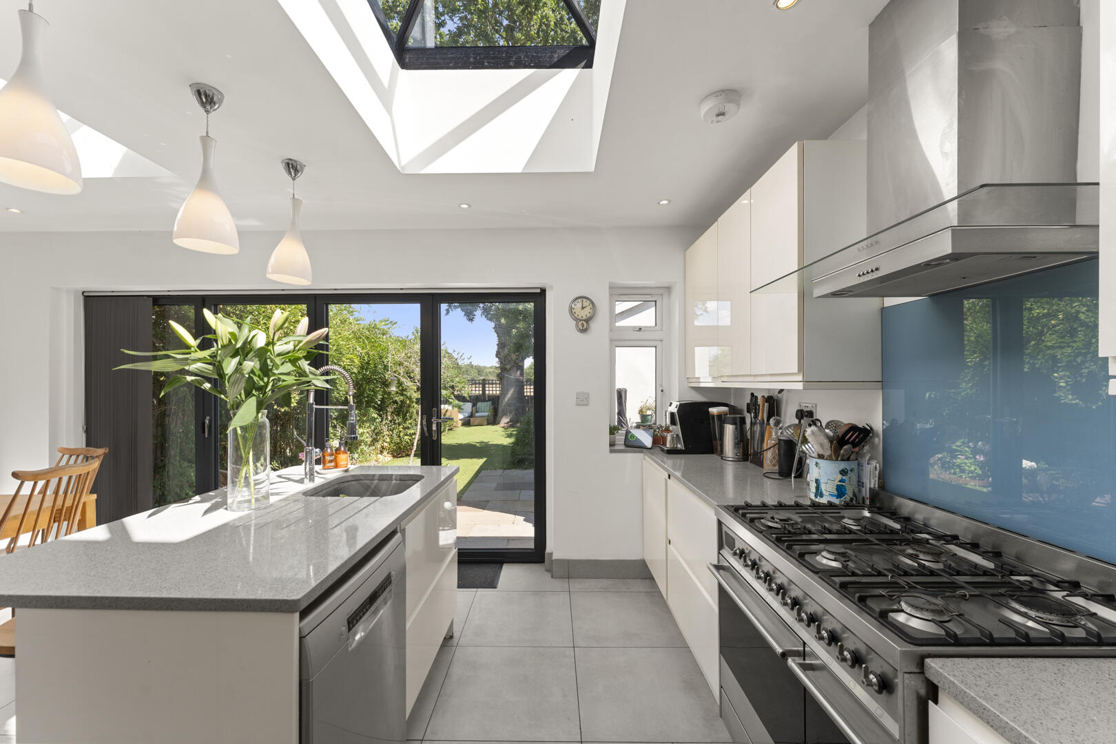 Bright, modern kitchen with a large island, stainless steel range, white cabinets, blue glass backsplash, and a skylight above a glass door to the garden outside.