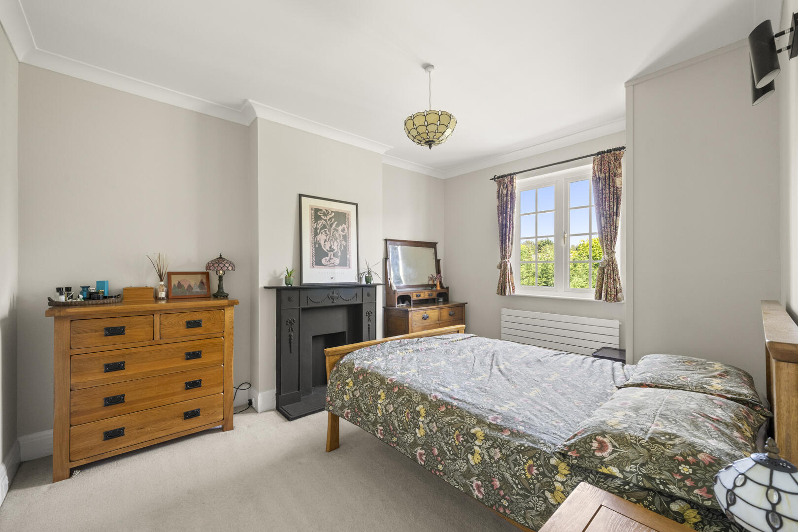 Cozy bedroom with a floral bedcover, wooden dresser on the left, and a black decorative fireplace mantel against pale walls; a window with floral curtains lets in sunlight on the right.