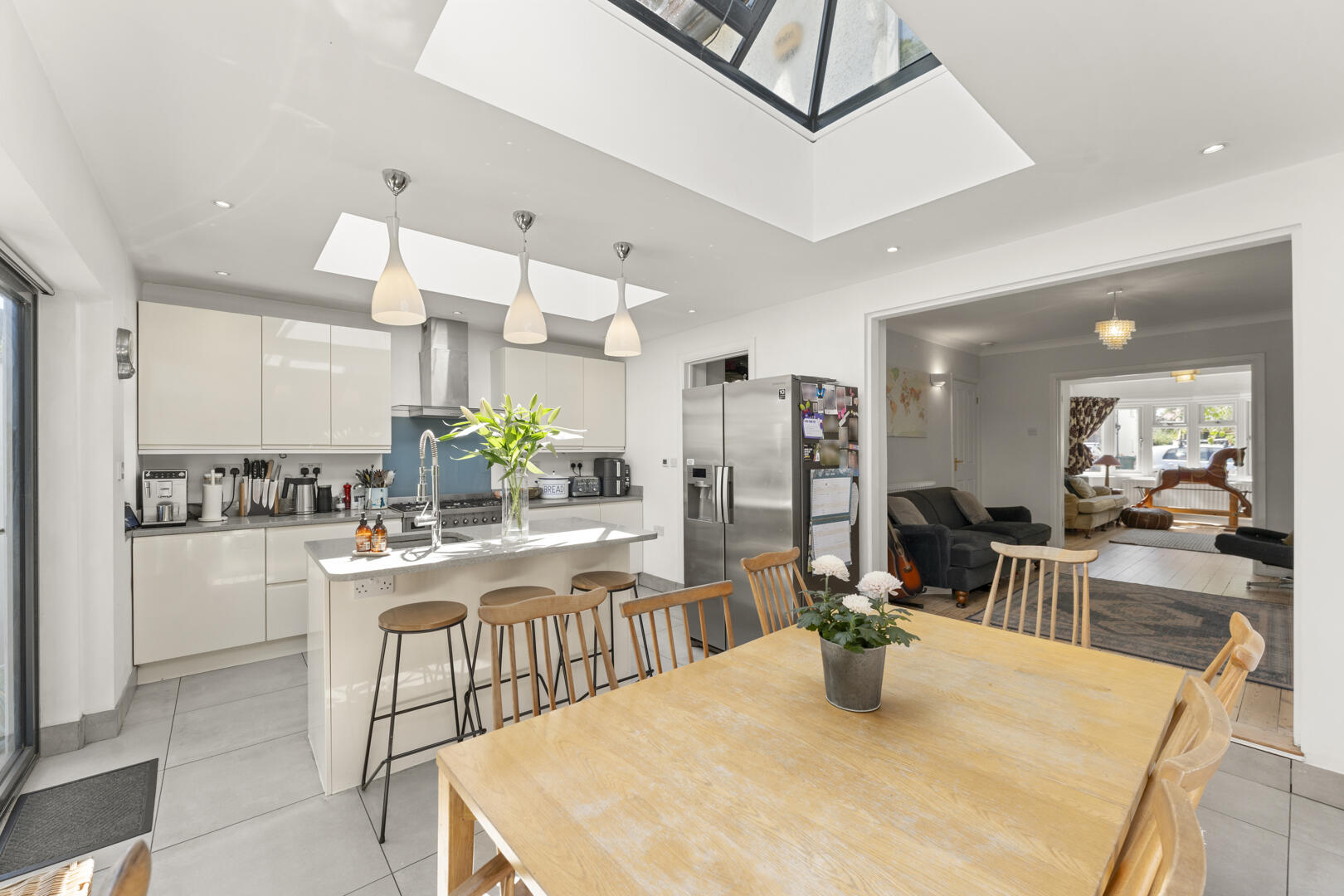 Bright open-plan kitchen with white cabinets, stainless fridge, pendant lights over the island, and a wooden dining table in the foreground.