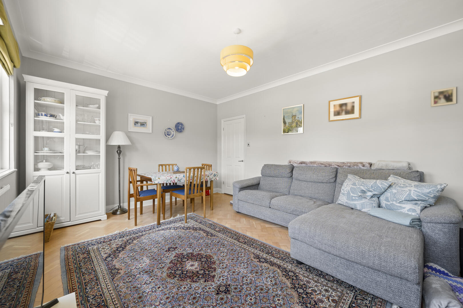 Living room with a gray sofa, patterned rug, and a white display cabinet by a dining table with four wooden chairs.