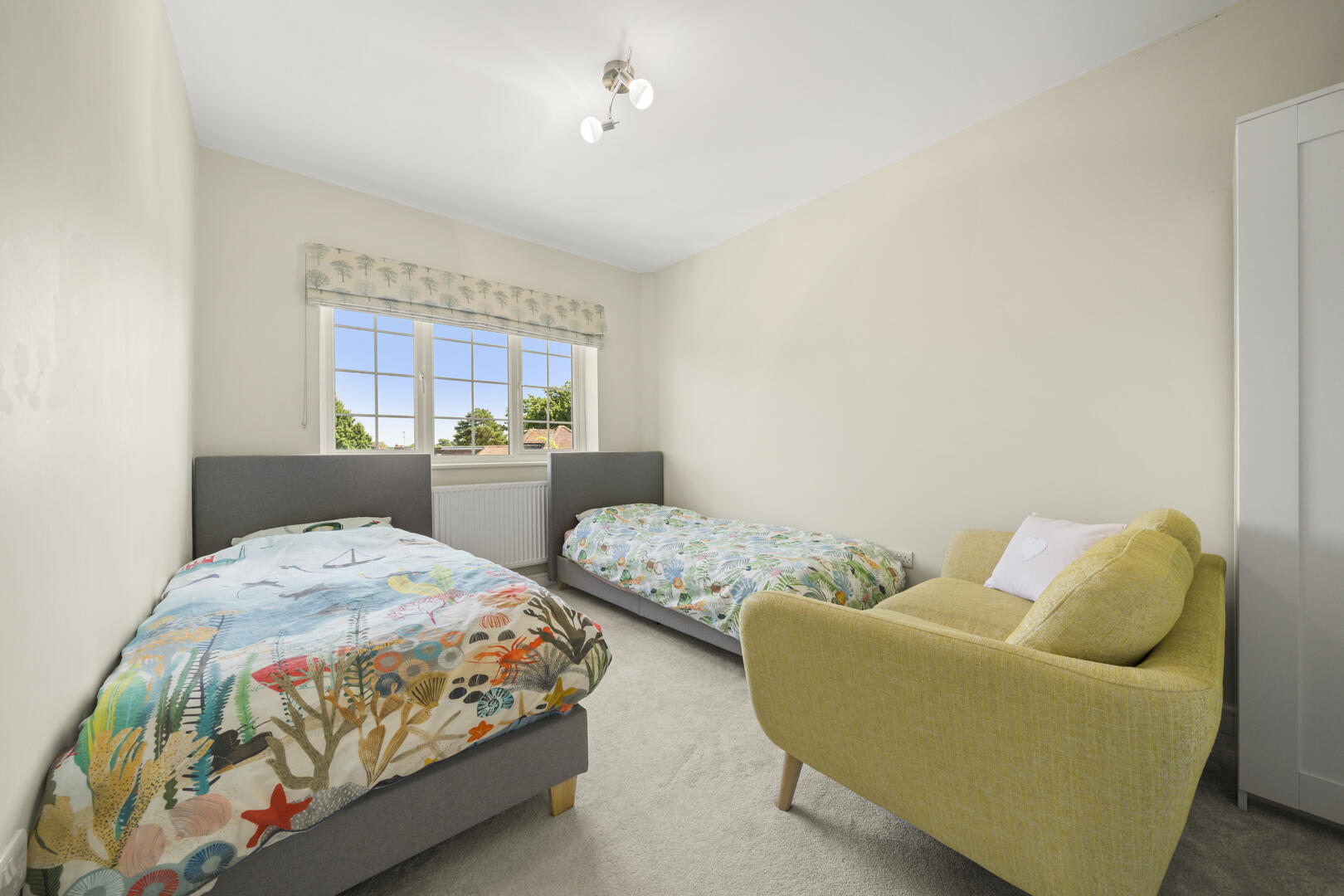 Twin bedroom with two gray upholstered beds, ocean-themed bedding, and a yellow lounge chair by a window.