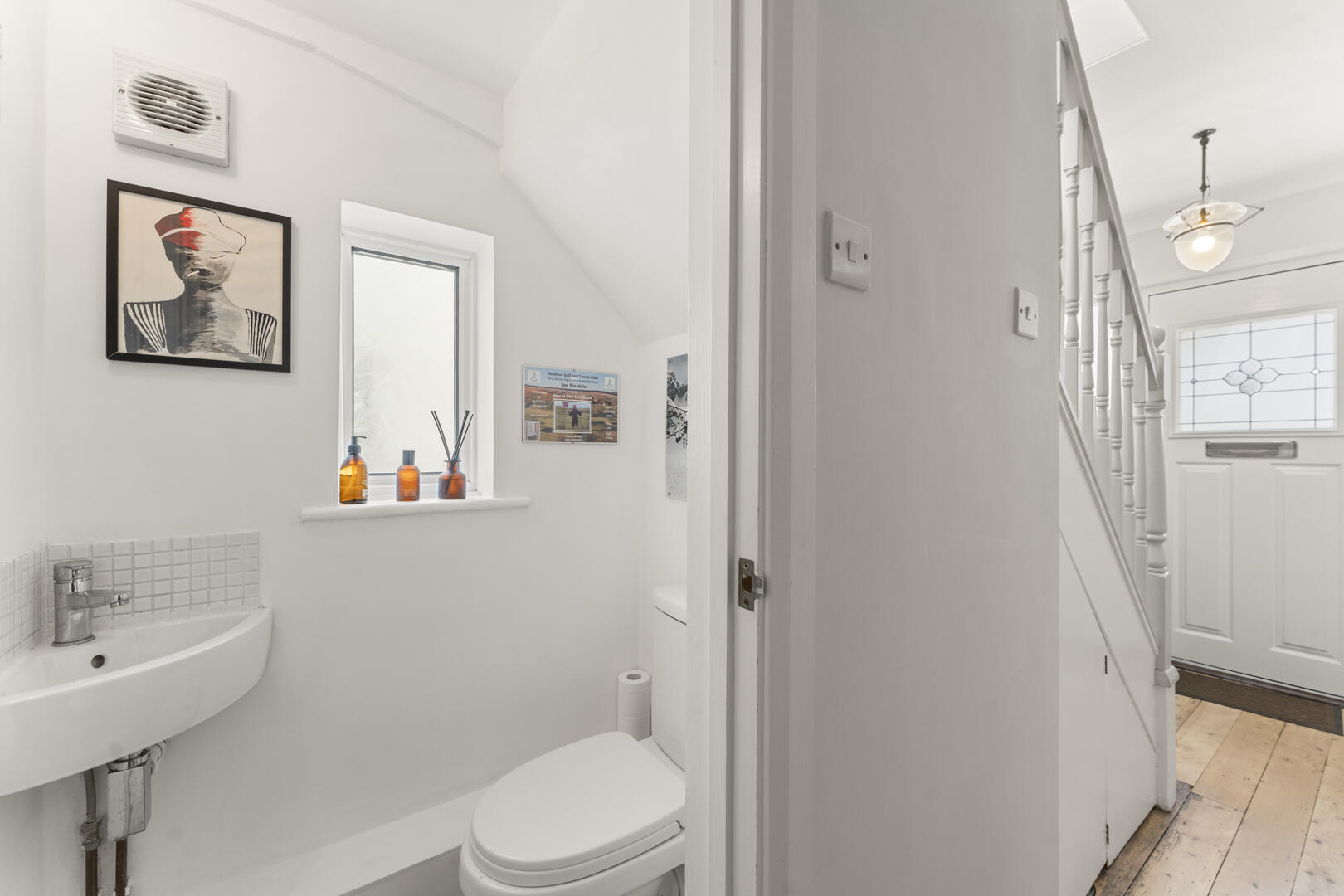 Bright white bathroom with a sink, toilet, and tub, a framed abstract portrait on the wall, and amber bottles on a window ledge.