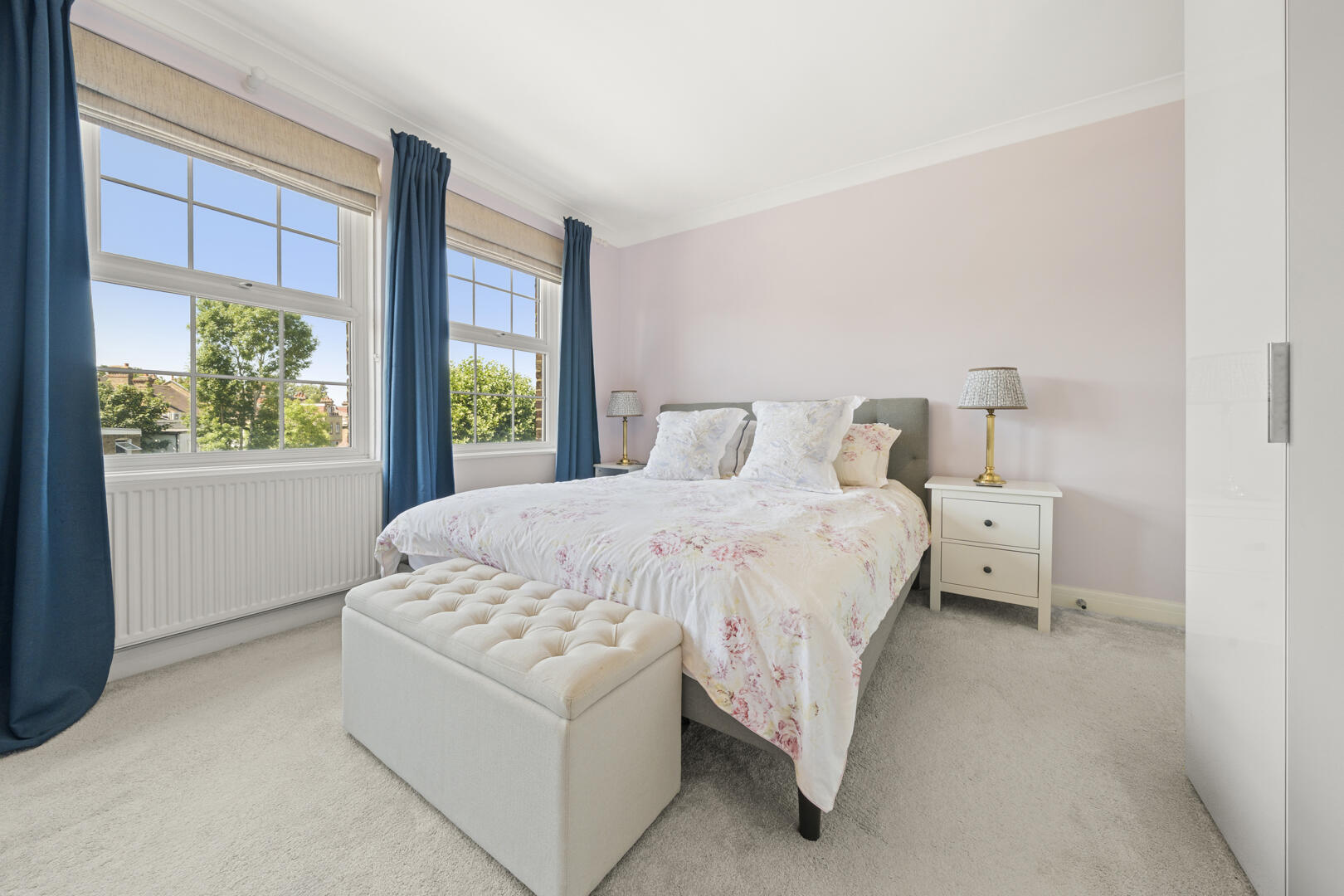 Bedroom with queen bed, floral duvet, tufted ottoman, and two nightstands with lamps under soft pink walls.