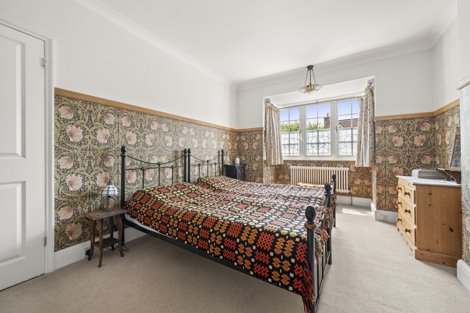 Cozy bedroom with a black metal double bed, a multicolored crocheted quilt, floral wallpaper, and a wooden dresser by a large window with curtains.