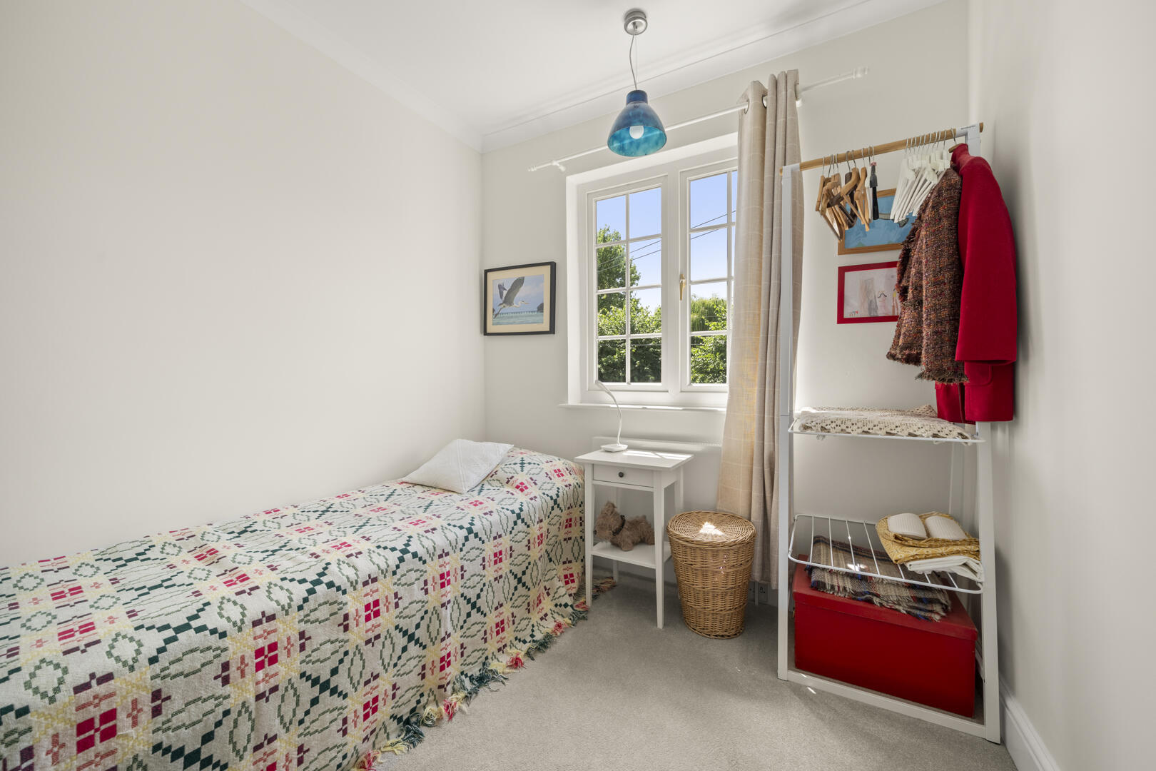 Bright small bedroom with a single bed in a colorful quilt, white nightstand, and a window with curtains; a wicker basket and clothing rack nearby.