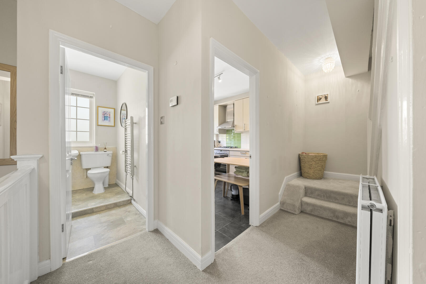 Bright hallway with a bathroom doorway to the left, a kitchen doorway ahead, and a wicker basket on the carpeted stairs to the right.