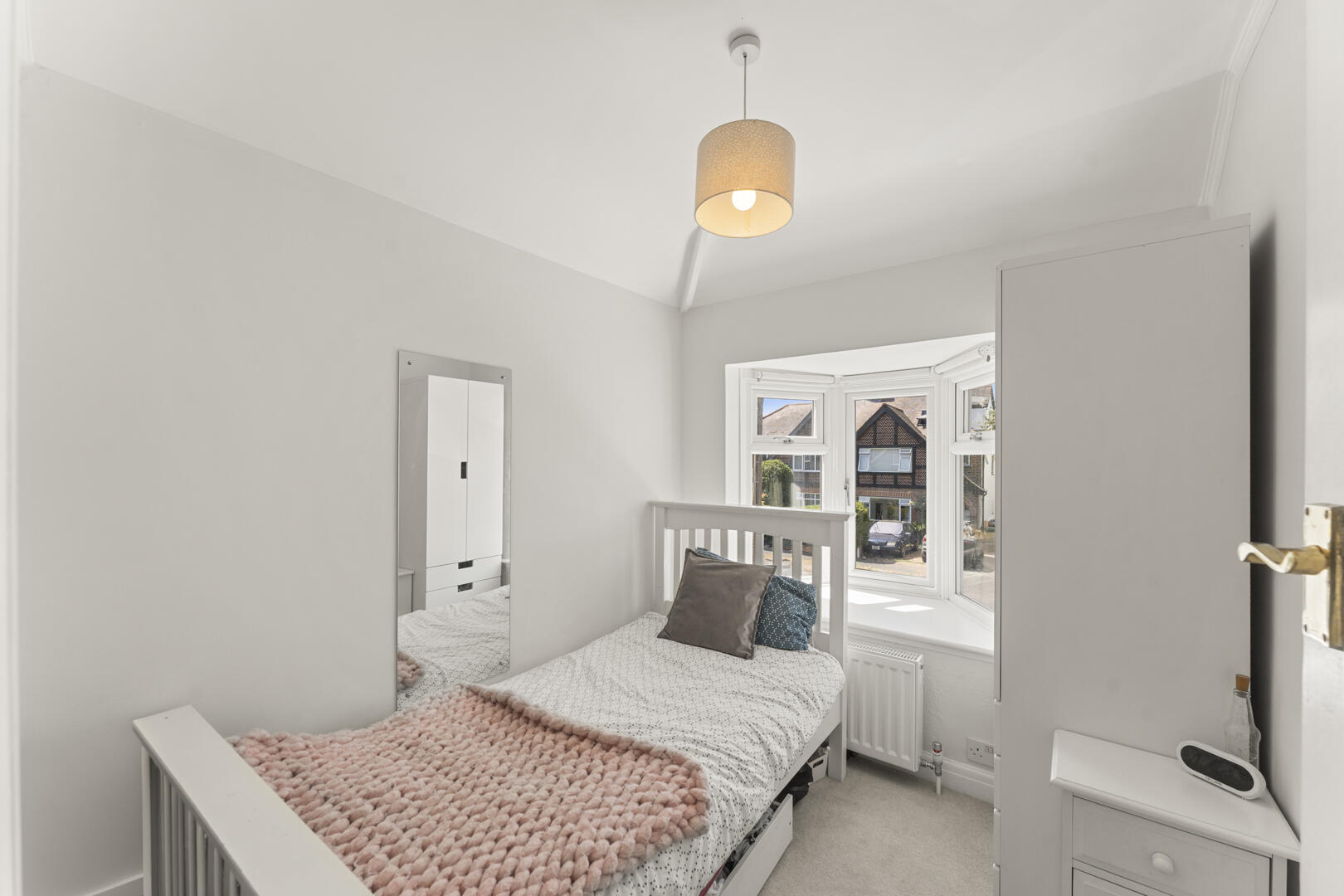 Small bright bedroom with a white wooden bed, pink chunky knit throw, and two decorative pillows facing a bay window that lets in daylight.