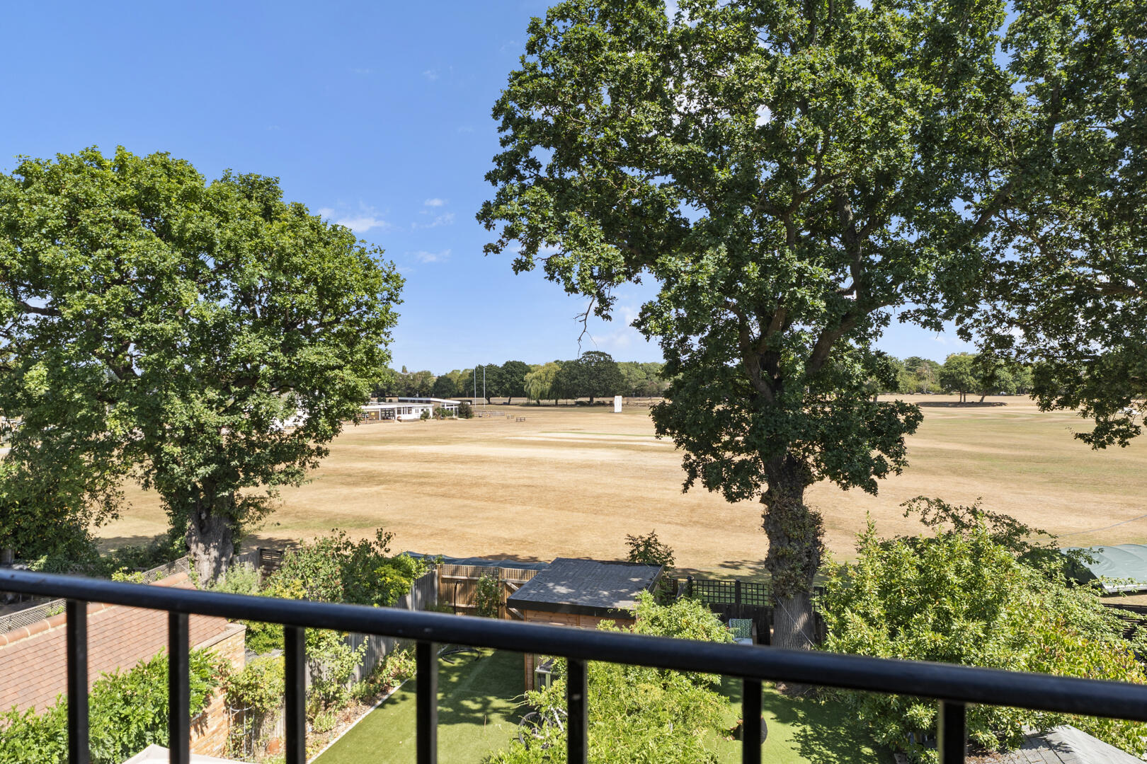 View from a balcony looking out over a park-like area with large green trees, a dry grass field, and a distant building under a blue sky.
