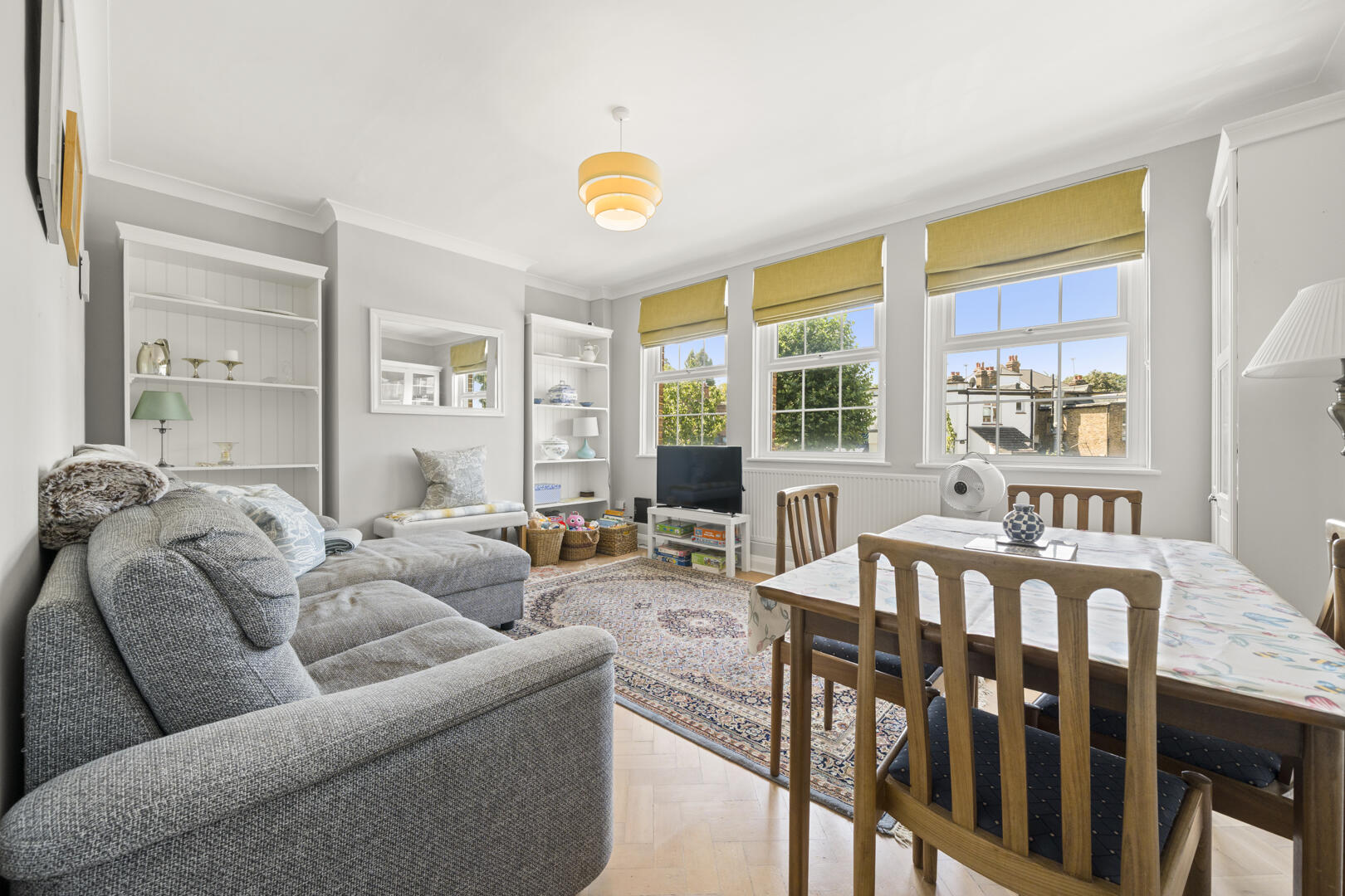 Bright living room with a gray sectional, white shelves, and a dining table near large windows with yellow Roman blinds outside light-wood floors. (Informative)