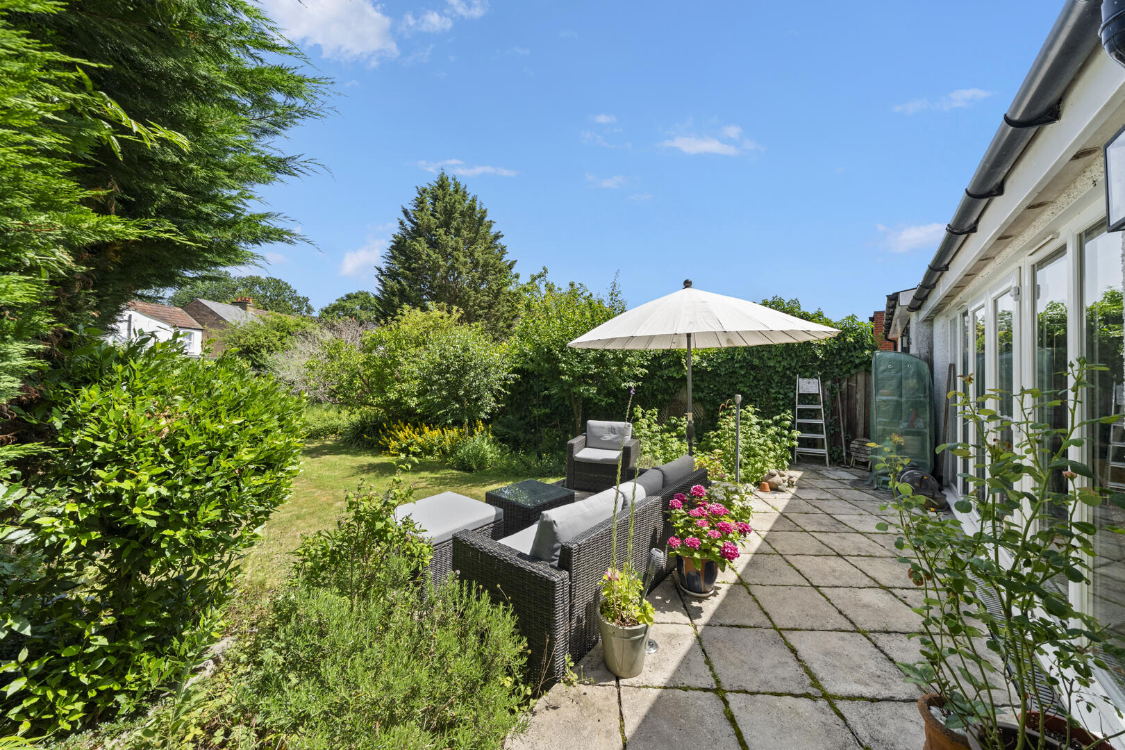 Sunny backyard patio with rattan seating under a large white umbrella and potted flowers beside a glass-walled house.