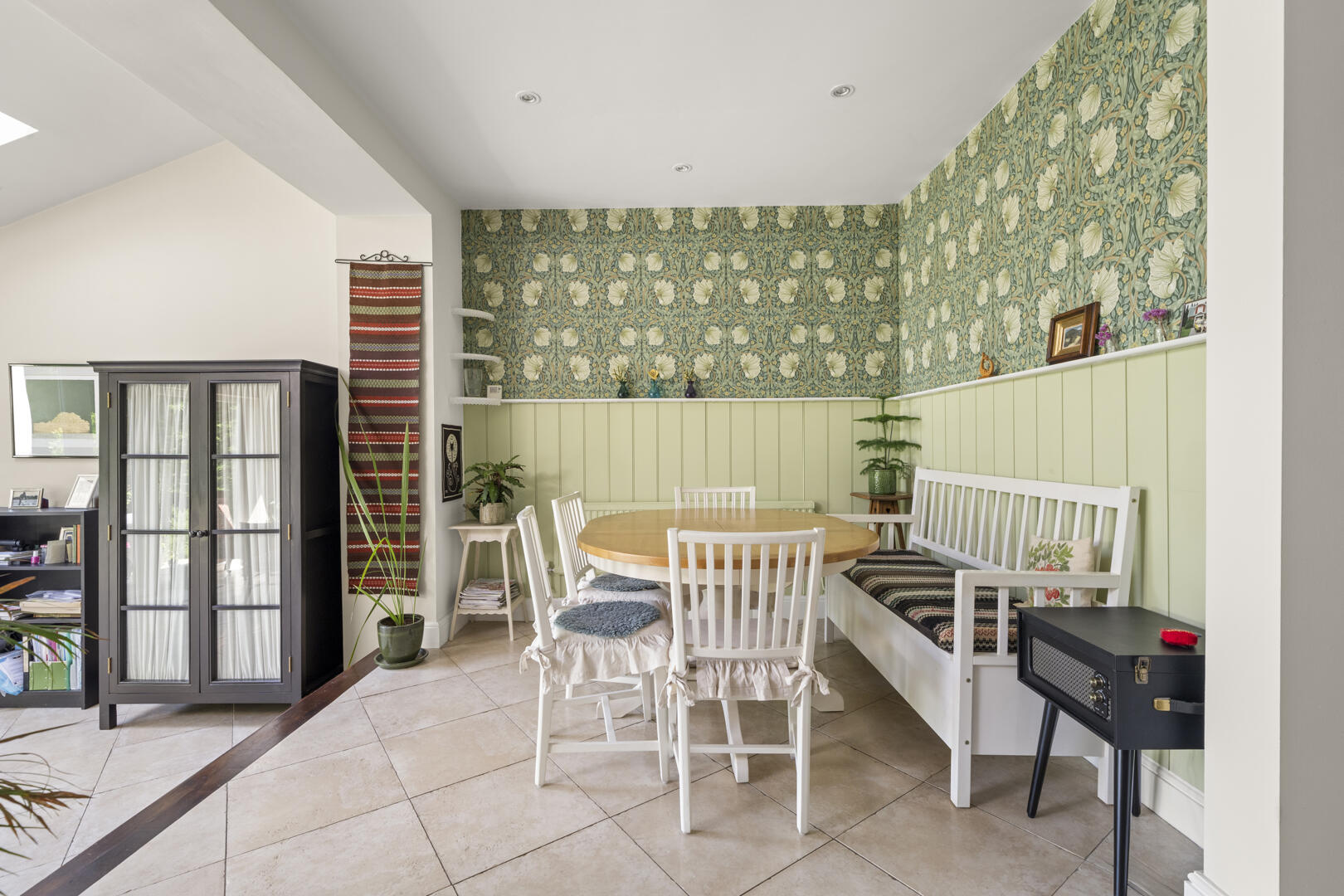 Round wooden table with white chairs and a white bench in a pastel-green dining nook, olive-green botanical wallpaper above pale wainscoting.