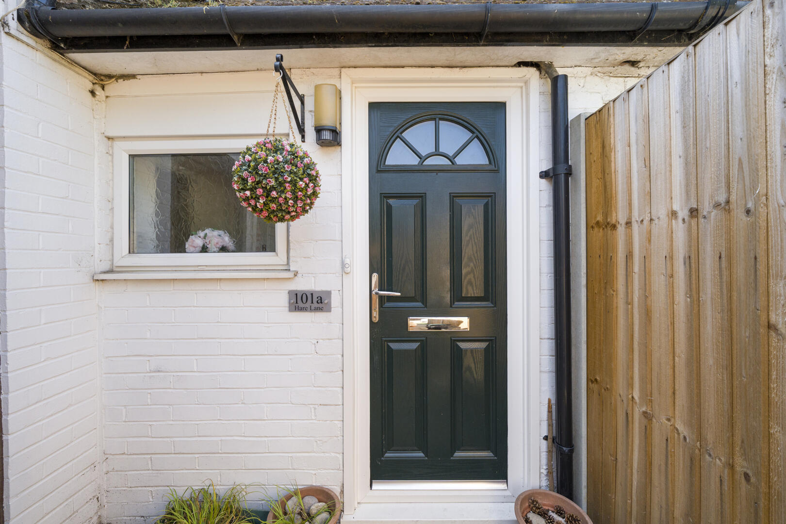 Dark green front door with an arched window, white trim, on a white brick wall; a hanging pink flower ball to the left of the doorway.