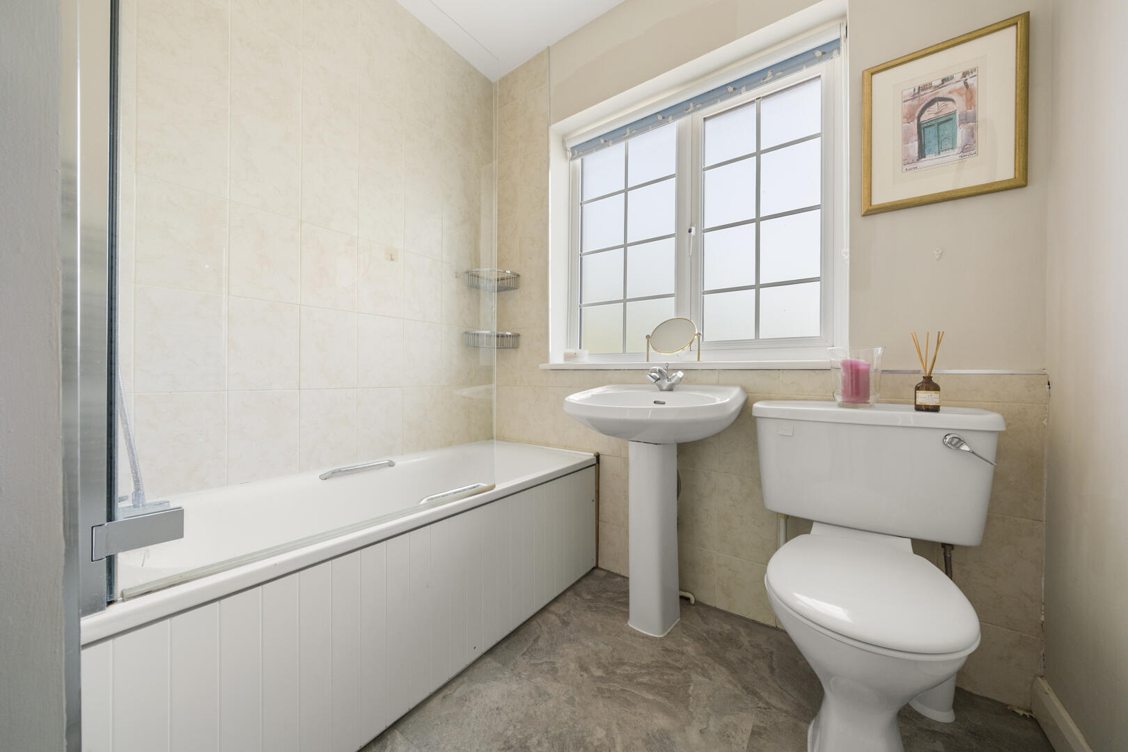Bright bathroom with a white bathtub and glass shower screen, beige tiled walls, pedestal sink, and toilet under a frosted window frame art on the wall.