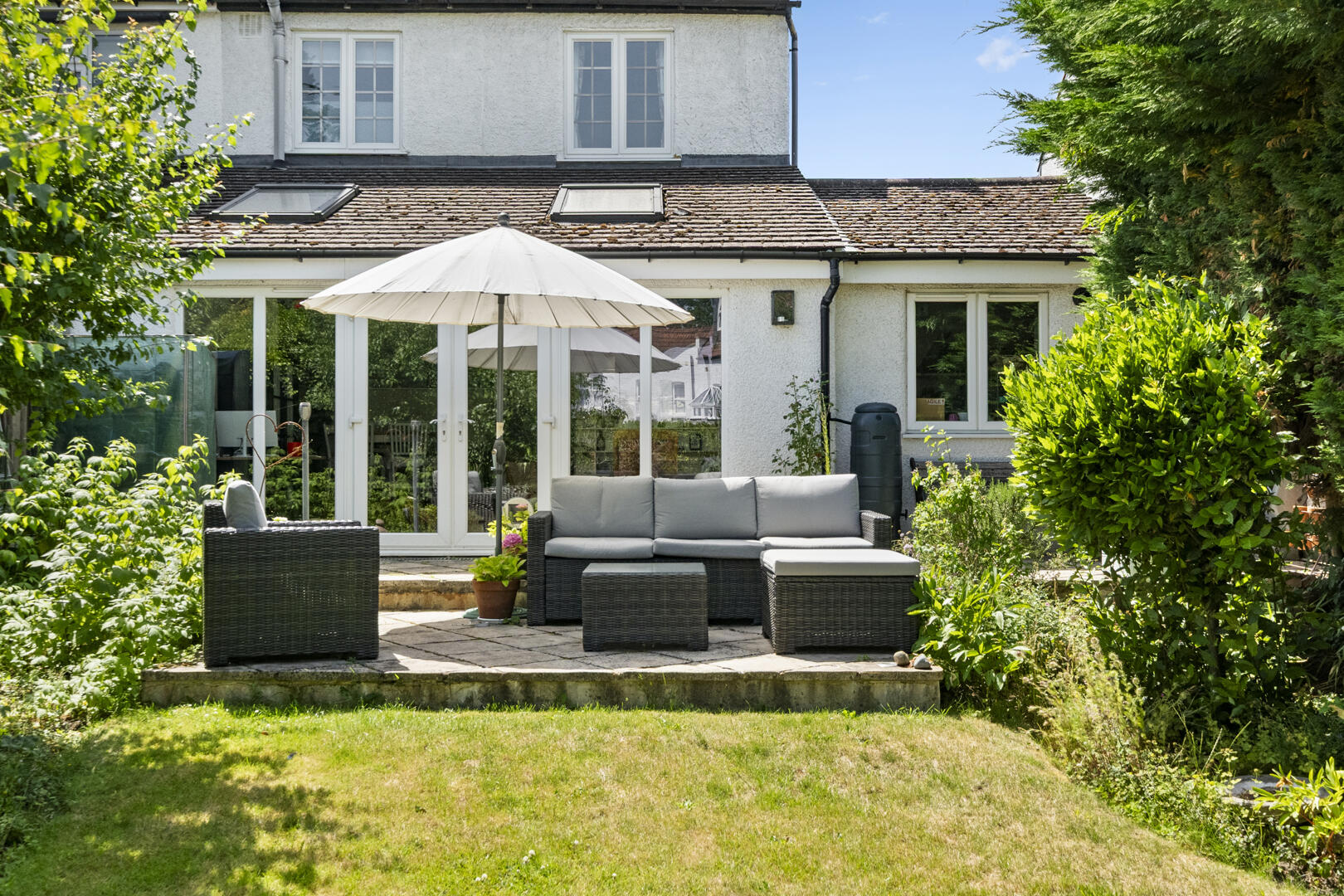 Backyard patio with a grey wicker sofa set under a white umbrella in front of a white house with glass doors.