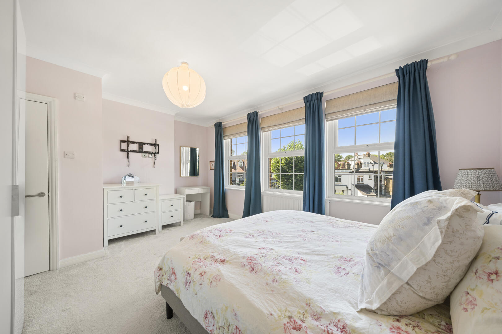 Bright pastel-pink bedroom with a floral quilt on the bed, white dresser and desk, and large windows with blue curtains outside rooftops.