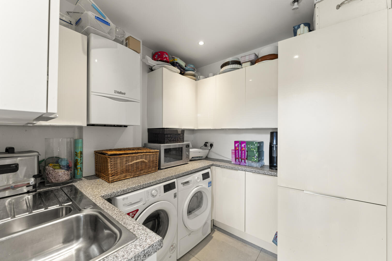 A compact laundry room with white cabinets, a sink, washing machines, and a microwave on a speckled countertop; laundry items stacked on top cabinets.