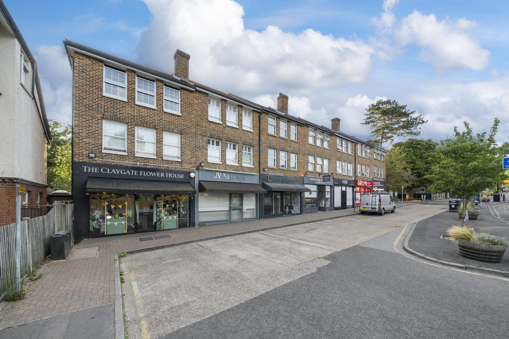 Row of brick storefronts along a quiet street, including The Claygate Flower House and JV Nails Spa under cloudy blue sky