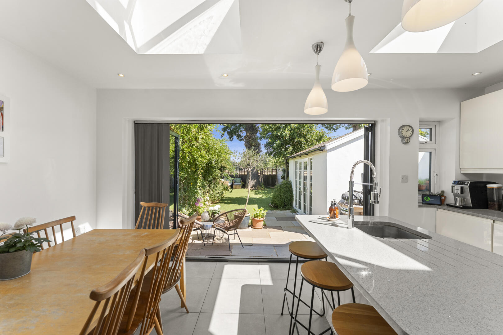Bright white kitchen with an island, bar stools, and a view open to a sunny garden patio and lawn outside.