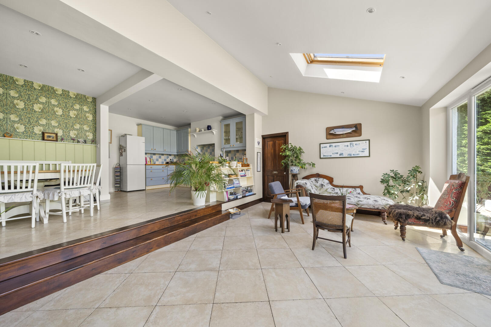 Bright open-plan living area with vintage furniture, tiled floor, and large windows; kitchen in background.