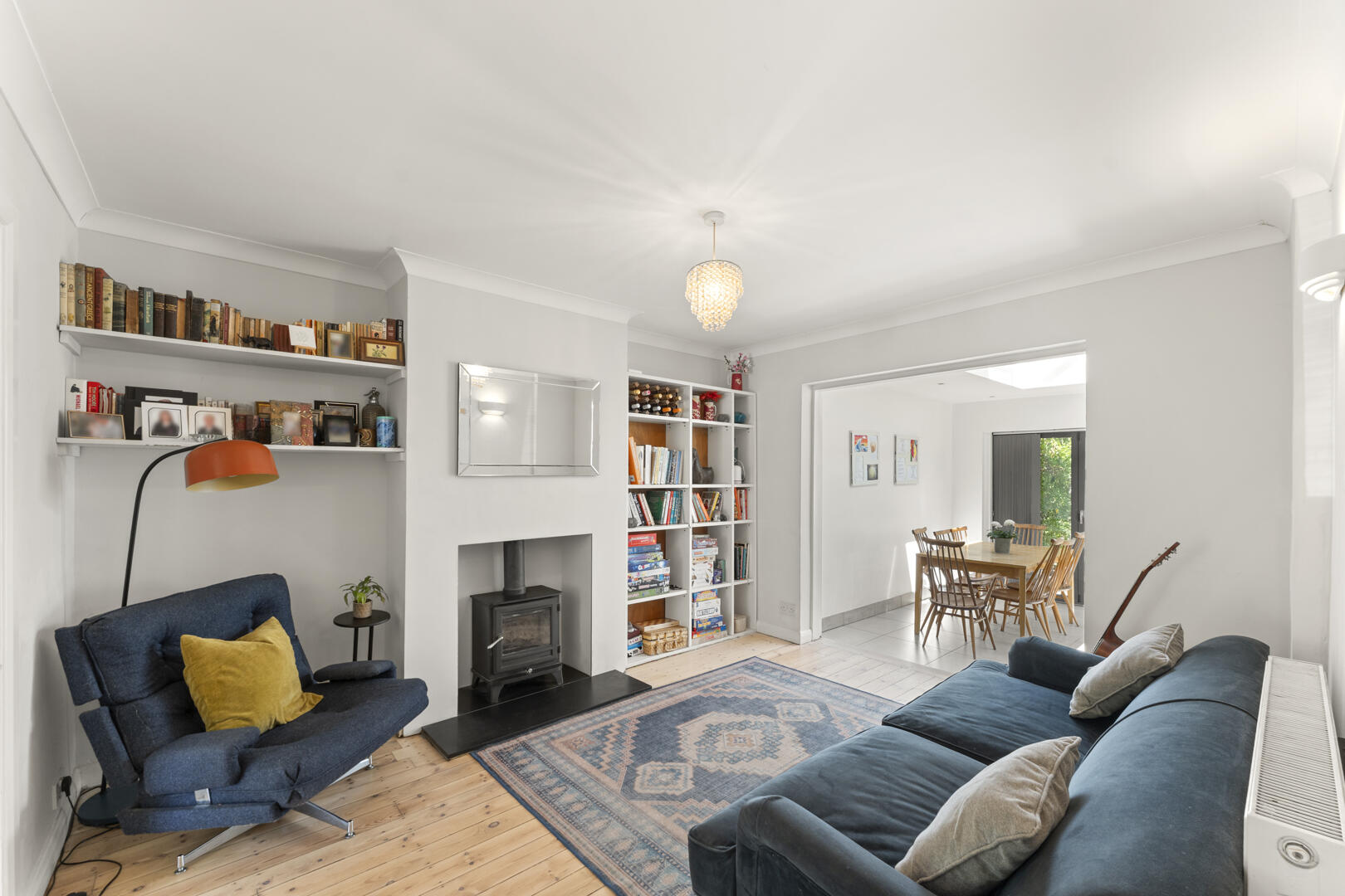 Bright living room with white walls, built-in bookshelves, a wood stove, and a blue sofa facing a rug-lined floor. A dining area is visible through an open doorway.