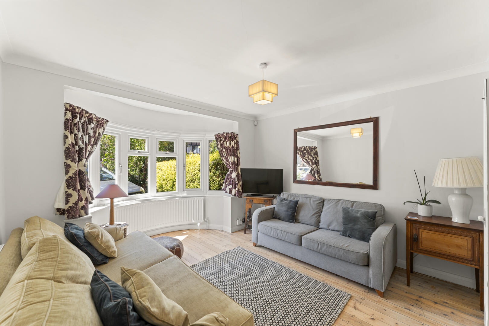 Bright living room with a bay window, two couches (beige and gray), patterned curtains, and a wall-mounted mirror above a wooden sideboard.