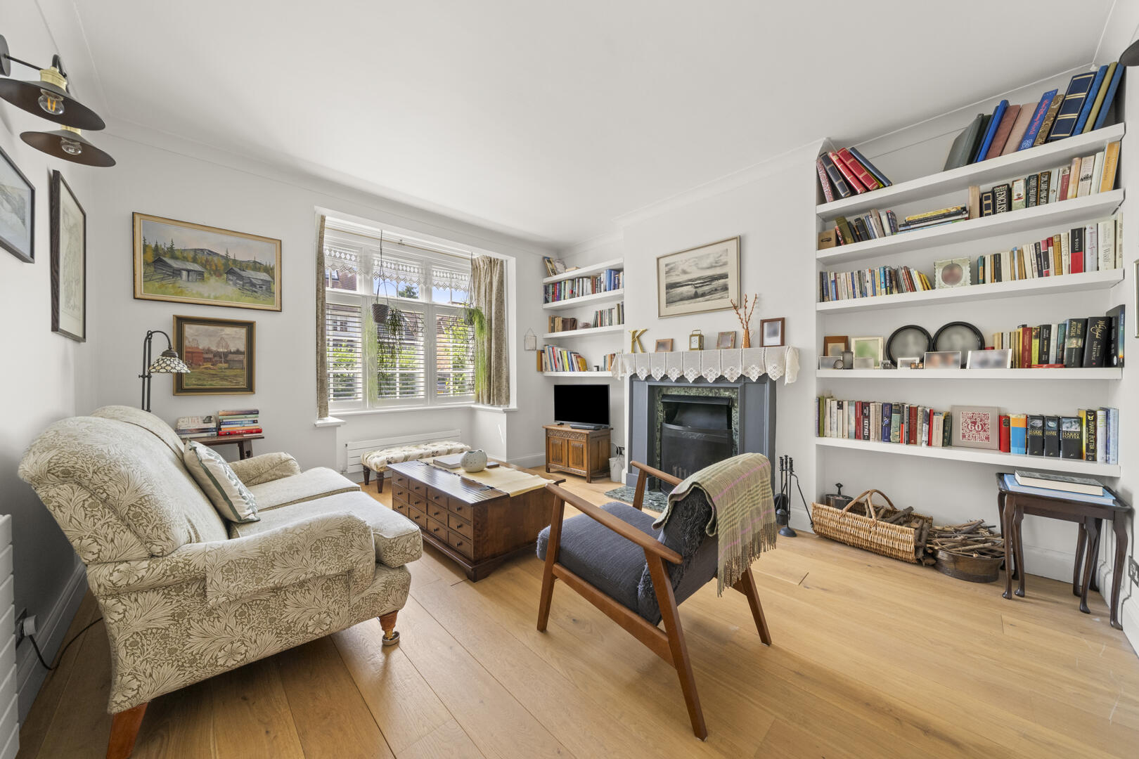 Bright living room with a fireplace, white built‑in bookshelves full of books, and two seating chairs around a wooden coffee table under a large window.