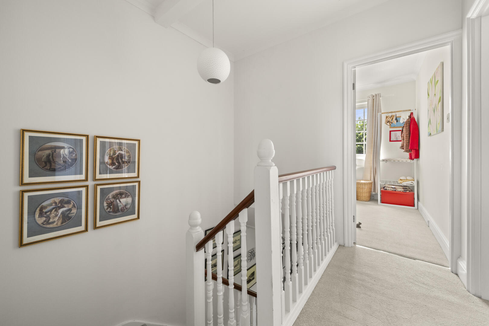 Upstairs hallway with white walls and a wooden handrail; four framed oval artworks on the left and a doorway to a dressing area with clothes and a red storage box.