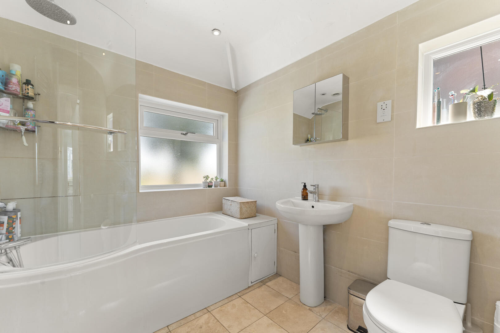 Modern beige-tiled bathroom with a white bathtub, glass shower screen, pedestal sink, mirrored cabinet, and a toilet; frosted window with small plants on the sill.