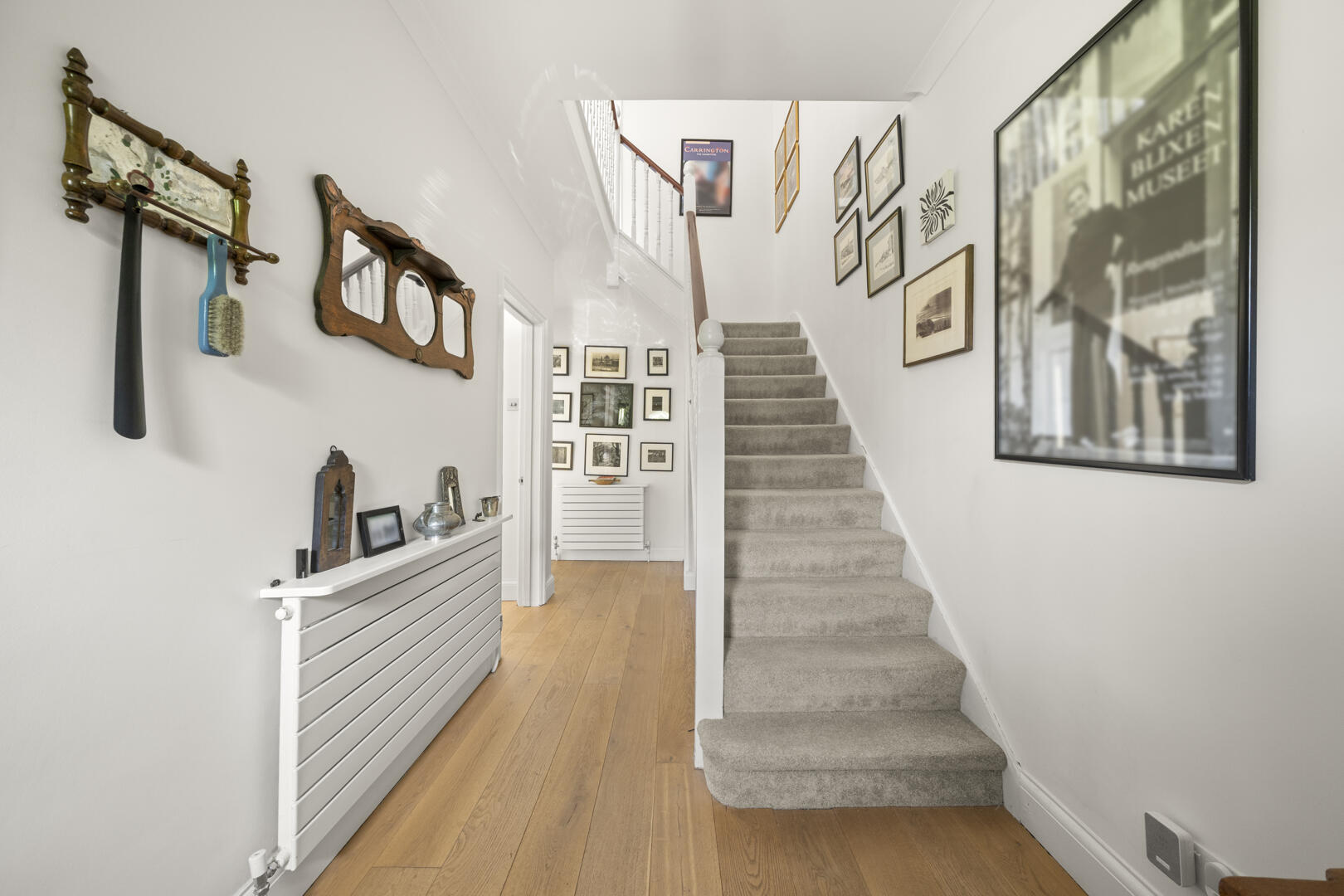 Bright hallway with light wood floor, white walls, and a carpeted staircase on the right; gallery wall of framed art along the stairs.