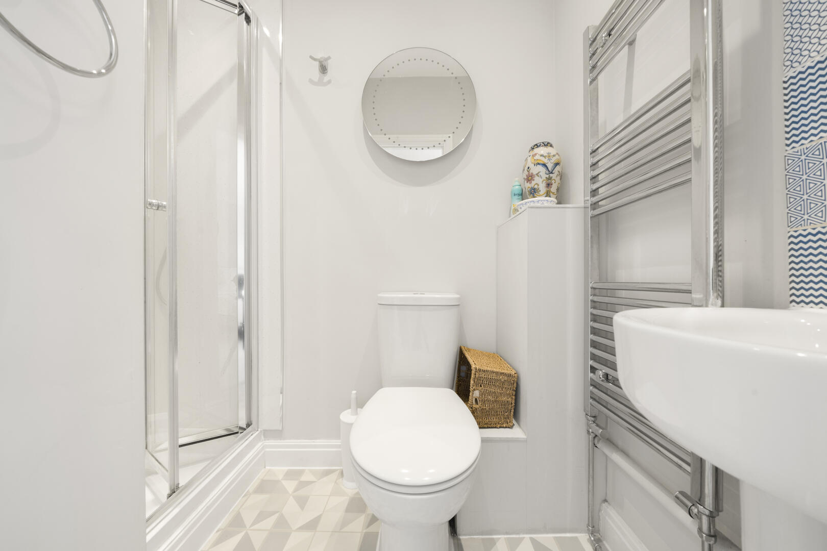 Small white bathroom with a glass shower, toilet, round wall mirror, wicker basket, and chrome towel rack on the right side.